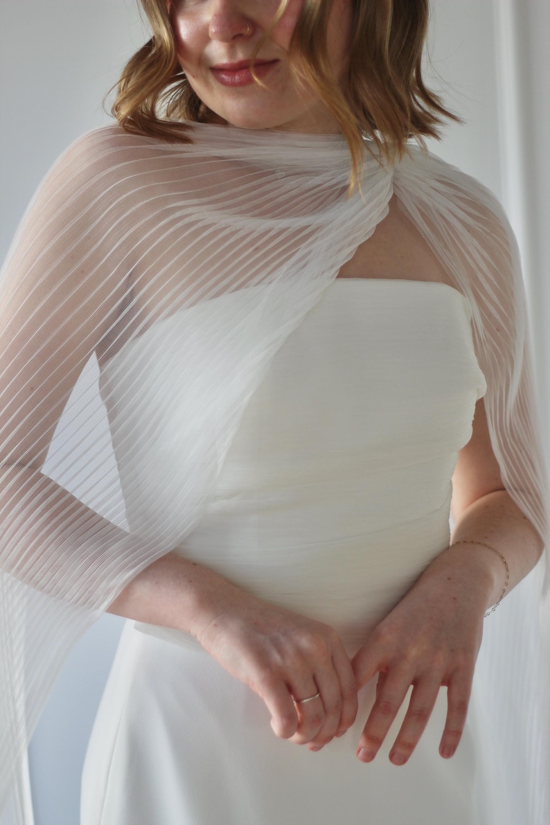 May include: A white, strapless wedding dress with a sheer, pleated wrap. The wrap drapes over the shoulders and arms. The dress is a simple, elegant design, perfect for a wedding or special occasion.