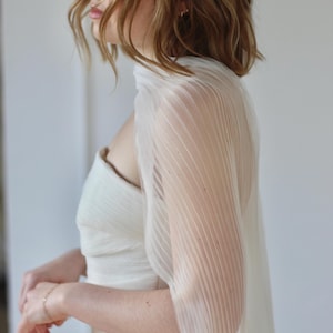 May include: A white, pleated, sheer bridal cape draped over a strapless wedding dress. The cape has vertical pleats and a flowing design. The dress is a simple, elegant style. The overall look is soft and romantic.
