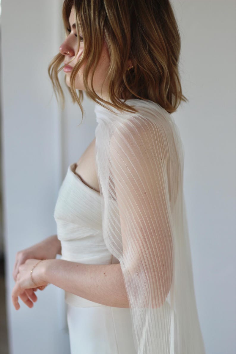 May include: A white, pleated, sheer bridal cape draped over a strapless wedding dress. The cape has vertical pleats and a flowing design. The dress is a simple, elegant style. The overall look is soft and romantic.