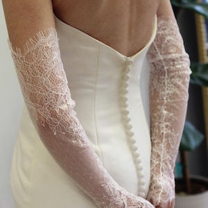May include: A white wedding dress with a lace overlay on the sleeves. The dress has a fitted bodice and a flowing skirt. The back of the dress features a row of buttons.