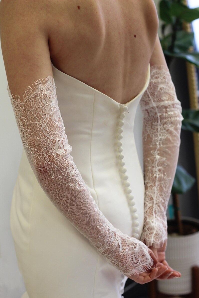 May include: A white wedding dress with a lace overlay on the sleeves. The dress has a fitted bodice and a flowing skirt. The back of the dress features a row of buttons.