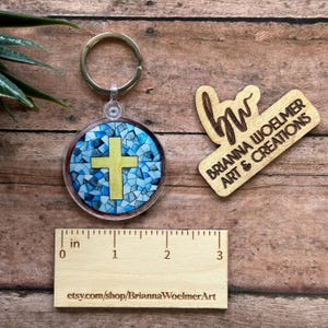 May include: A blue and yellow stained glass cross keychain with a wooden tag that reads "Briana Woelmer Art & Creations".
