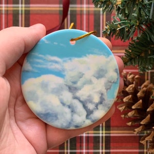 May include: A round ceramic ornament with a blue sky and white clouds painted on it. The ornament has a gold string attached to it.