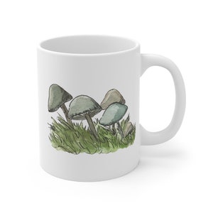 Pode incluir: Caneca de cer&acirc;mica branca com uma ilustra&ccedil;&atilde;o desenhada &agrave; m&atilde;o de cogumelos em tons de verde e azul, cercados por grama verde.