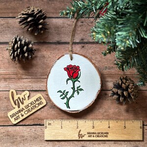 May include: A round wooden ornament with a white background and a red rose painted on it. The ornament is about 3 inches in diameter and has a twine string for hanging.