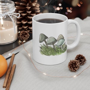 Pode incluir: Caneca de cer&acirc;mica branca com al&ccedil;a preta. A caneca tem uma ilustra&ccedil;&atilde;o em aquarela de cogumelos em tons de verde e cinza sobre um fundo branco.