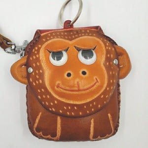 May include: A brown leather coin purse shaped like a monkey's face, featuring large white eyes and a smiling mouth. The purse has a key ring and a brown leather strap. The monkey's face is detailed with stitched accents.