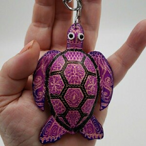 May include: A purple turtle keychain with a silver keyring. The turtle has a detailed design with black outlines and gold accents. The keychain is held in a hand.