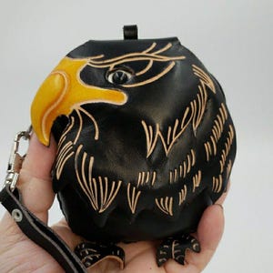 May include: A black leather pouch shaped like an eagle head with a bright yellow beak and carved feather details. The pouch features a dark eye, a metal clasp, and a black leather wrist strap attached to the side for carrying convenience.