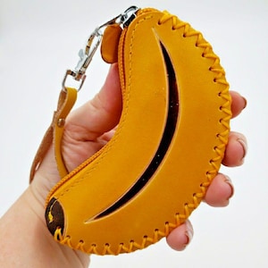 May include: A yellow banana-shaped leather coin purse with a black stripe detail. The purse has a zipper closure and a silver-tone clasp with a wrist strap. The edges are stitched with yellow thread.