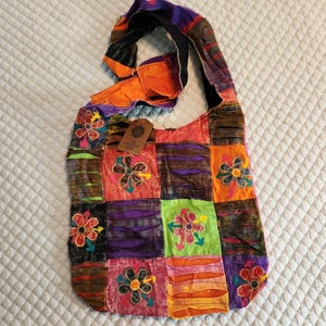 May include: A hobo bag featuring a patchwork design of vibrant, multi-colored fabric squares in shades of orange, purple, green, and black. The bag is decorated with embroidered floral motifs and includes a brown rectangular tag with text.