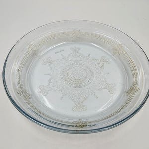 May include: A clear glass plate featuring an embossed central medallion with intricate scrollwork and floral patterns. The rim is decorated with a repeating geometric border and symmetrical flourishes, creating a textured surface design.