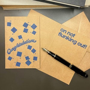 May include: Two brown cards with blue text. The first card has a pattern of blue graduation caps and the text "Congratulations". The second card has the text "on not flunking out!" A black and gold pen is lying on top of the cards.