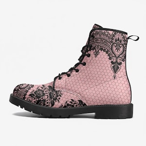 Puede incluir: Un botín con cordones que presenta una base rosa claro con un patrón de malla hexagonal negra y detalles intrincados de encaje floral negro en la puntera y el cuello. La bota tiene cordones negros, ojales negros y una suela de goma gruesa.