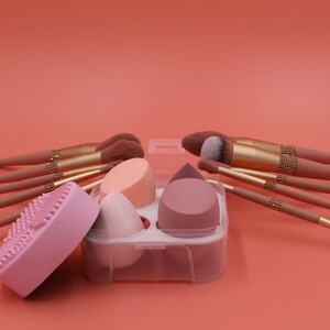 Puede incluir: Un conjunto de brochas de maquillaje y esponjas en un contenedor de pl&aacute;stico transparente. Las brochas tienen mangos rosas con detalles dorados y las esponjas son rosas, melocot&oacute;n y beige.