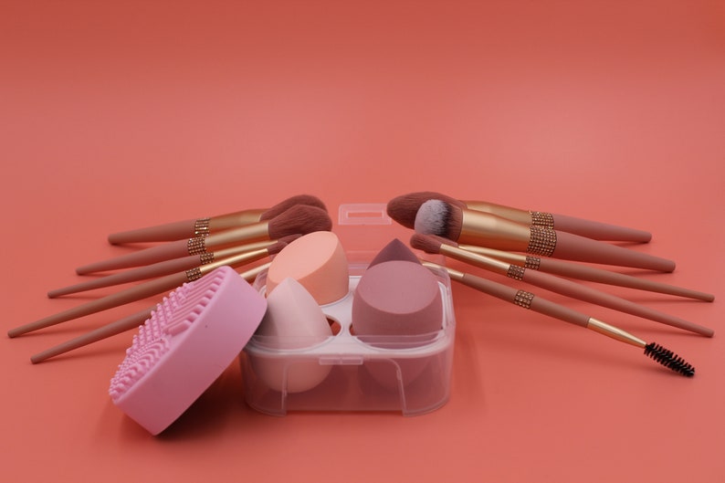 Puede incluir: Un conjunto de brochas de maquillaje y esponjas en un contenedor de pl&aacute;stico transparente. Las brochas tienen mangos rosas con detalles dorados y las esponjas son rosas, melocot&oacute;n y beige.