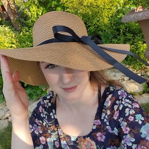 May include: A brown straw sun hat with a wide brim and a black ribbon tied in a bow. The hat is worn by a person with long brown hair.