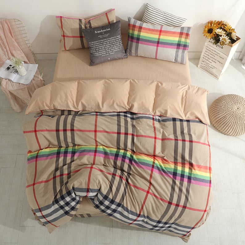 May include: Beige and brown plaid bedding with a rainbow stripe. The bedding is on a bed with two pillows, one with a rainbow stripe and one with a gray pillow with the word "Scandinavian" printed on it.