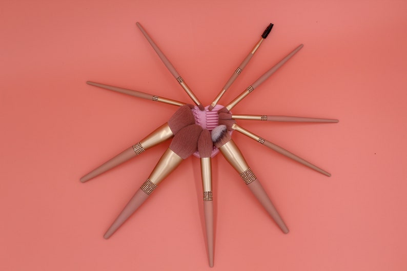 Puede incluir: Un conjunto de ocho pinceles de maquillaje con mangos de oro rosa y cerdas rosadas. Los pinceles est&aacute;n dispuestos en forma de estrella sobre un fondo coral.