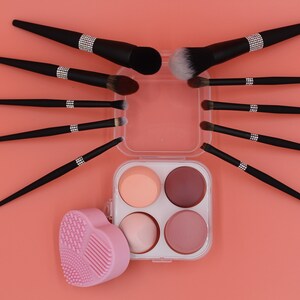 Puede incluir: Un conjunto de pinceles de maquillaje negros con detalles de pedrer&iacute;a, dispuestos en forma de estrella alrededor de una caja de pl&aacute;stico transparente con cuatro esponjas redondas. La caja tiene una esponja rosa en forma de coraz&oacute;n en el lado izquierdo.