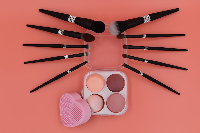 Puede incluir: Un conjunto de pinceles de maquillaje negros con detalles de pedrer&iacute;a, dispuestos en forma de estrella alrededor de una caja de pl&aacute;stico transparente con cuatro esponjas redondas. La caja tiene una esponja rosa en forma de coraz&oacute;n en el lado izquierdo.