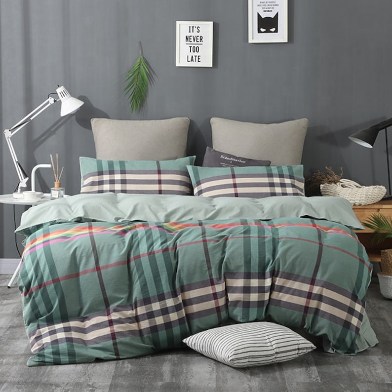May include: A green and white plaid duvet cover with a colorful stripe detail. The duvet cover is on a bed with two pillows, one with a plaid pattern and the other with a solid color. The bed is in a bedroom with a gray wall and a lamp on a side table.