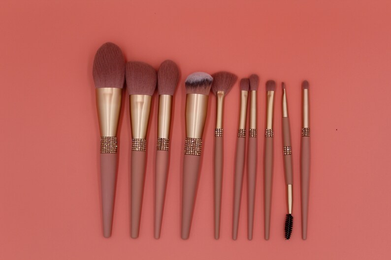 Puede incluir: Conjunto de diez pinceles de maquillaje rosa y dorado con un detalle de pedrer&iacute;a en los mangos. Los pinceles tienen diferentes formas y tama&ntilde;os para diversas aplicaciones de maquillaje.