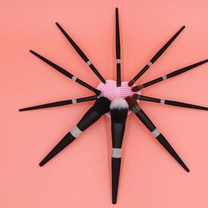 Puede incluir: Un conjunto de pinceles de maquillaje negros con detalles de pedrer&iacute;a dispuestos en forma de estrella sobre un fondo rosa. Los pinceles est&aacute;n dispuestos alrededor de una esponja de maquillaje rosa en forma de coraz&oacute;n.