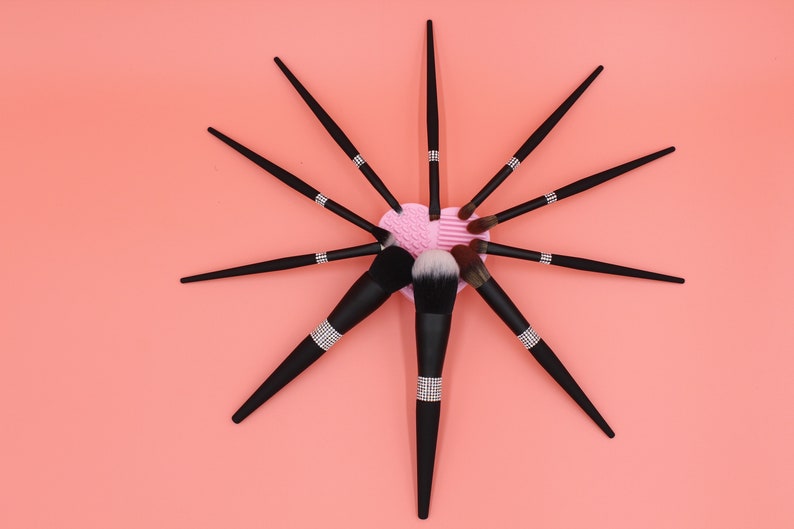 Puede incluir: Un conjunto de pinceles de maquillaje negros con detalles de pedrer&iacute;a dispuestos en forma de estrella sobre un fondo rosa. Los pinceles est&aacute;n dispuestos alrededor de una esponja de maquillaje rosa en forma de coraz&oacute;n.