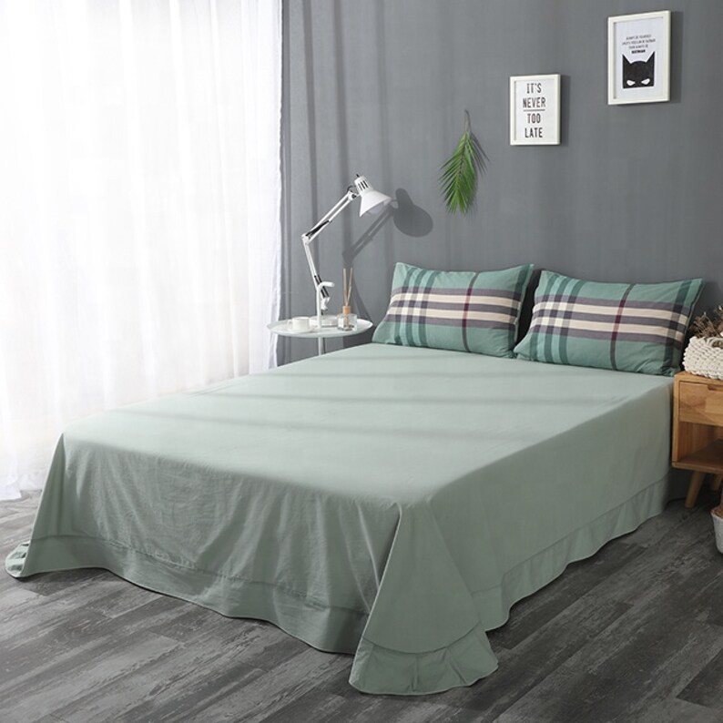May include: A green bed sheet with a white border and two plaid pillows. The pillows have a black, white, and green plaid pattern. The bed is in a bedroom with a gray wall and a white lamp.