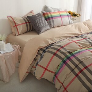 May include: Beige and brown plaid bedding with a rainbow stripe detail. The bedding is arranged on a bed with several pillows, including a gray pillow with text and a striped pillow.
