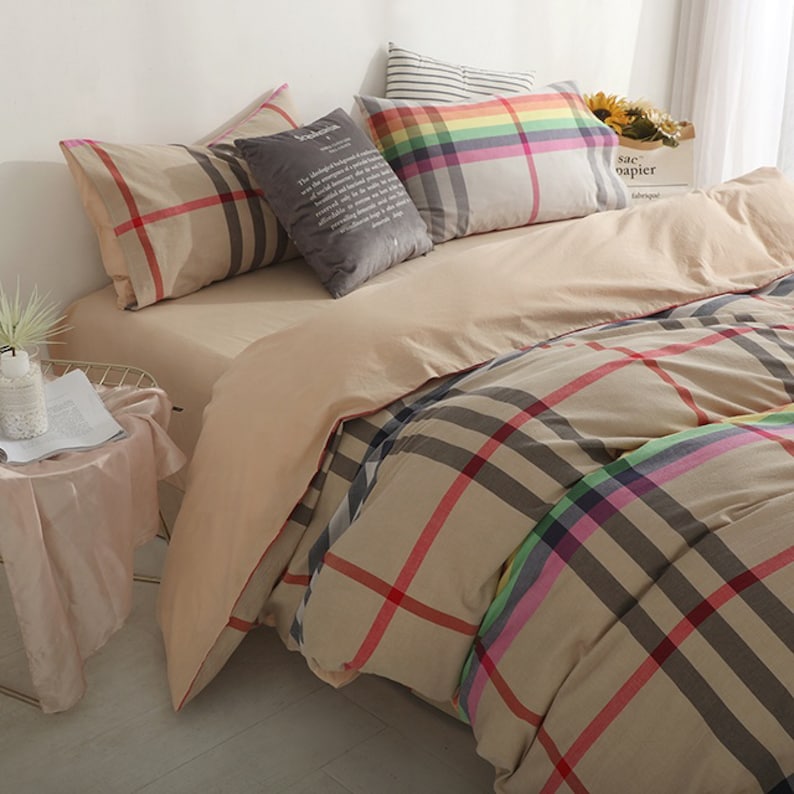 May include: Beige and brown plaid bedding with a rainbow stripe detail. The bedding is arranged on a bed with several pillows, including a gray pillow with text and a striped pillow.