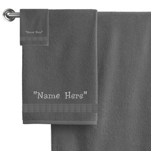 May include: Two gray towels hanging on a silver towel rack. The larger towel has the text "Name Here" embroidered on it. The smaller towel has the same text embroidered on it.