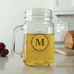 May include: A clear glass mason jar mug with a black handle. The mug has a black and white design with a wreath and the letter "M" in the center. The text "Natalia & Andrew Micalson September 14, 2023" is printed on the mug.