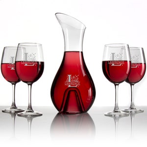 Puede incluir: Una jarra de vino de cristal transparente y cuatro copas de vino a juego llenas de vino tinto. Cada copa y la jarra están grabadas con el nombre "Lawrence" y una inicial "L" adornada. Las copas de vino tienen tallos delgados.