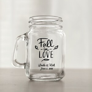Puede incluir: Una taza de vidrio transparente tipo tarro de albañil con un asa negra. La taza tiene una calcomanía de vinilo negro que dice "Fall -in- LOVE" con un diseño de hoja a cada lado del texto. Debajo del texto se lee "Linda & Rick June 8, 2026".