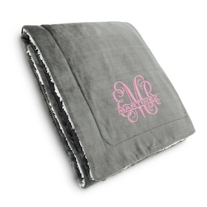 May include: A gray fleece throw blanket with a white sherpa lining. The blanket has a pink monogram with the name "MARTINEZ" embroidered on it.
