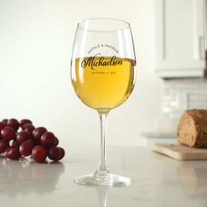 May include: A clear glass wine glass with a personalized inscription that reads "Natalie & Andrew Michaelson September 14, 2018". The glass is filled with white wine and is sitting on a white countertop.