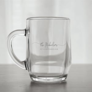 May include: Clear glass mug with a handle. The mug has the text "the Michaelsons" in a cursive font, with "EST 14 SEPTEMBER 2028" below it. The mug is sitting on a light-colored surface.