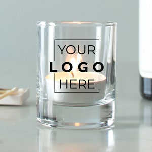 May include: A clear glass candle holder with a white tea light candle inside. The holder has the text "YOUR LOGO HERE" printed on the side.