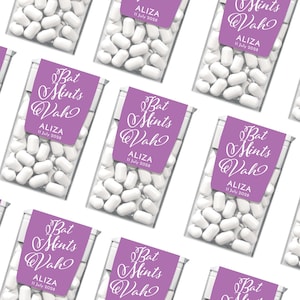 May include: Purple and white favor boxes filled with white mints. The boxes have a label with the text "Bat Mints Wah" and the name "ALIZA" with the date "11 July 2028".