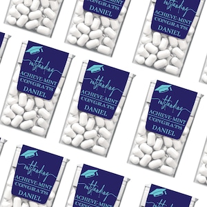 May include: Multiple clear plastic boxes filled with white mints. Each box has a navy blue label with the text "outstanding", "ACHIEVE-MINT CONGRATS DANIEL", and a graduation cap graphic. The boxes are arranged on a white background.