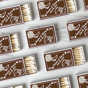 May include: Brown matchboxes with white text and illustrations. Each box is labelled "Sandra & Tommy July 8th 2029" with a heart and a drawing of a cowboy boot and flowers. Some boxes show the matches inside.