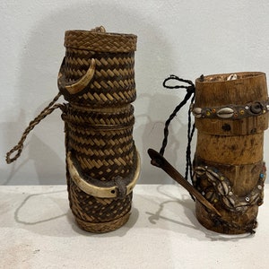 以下が含まれることがあります： 装飾的なアクセントが付いた2つの織り容器。左側の容器は、編まれたわらでできており、ハンドルが付いています。右側の容器は木でできており、上部に貝殻とビーズのバンドが付いています。