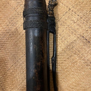 Puede incluir: Un recipiente de madera marrón oscuro con un asa de madera tallada y una parte superior decorativa. El recipiente está envuelto con un cordón negro y tiene un aspecto desgastado y envejecido.