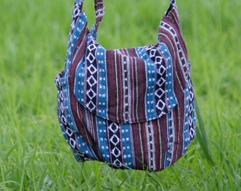 May include: A blue and brown patterned shoulder bag with a wooden handle. The bag is hanging from a person's hand in a field of green grass.