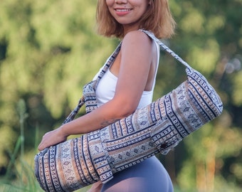 May include: A woman in a white tank top and blue yoga pants is carrying a patterned yoga mat bag over her shoulder. The bag has a long strap and is made of a woven fabric with brown, white, and black geometric designs.