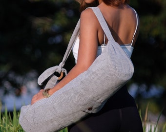 May include: A gray yoga mat bag with a long strap. The bag is made of a textured fabric and has a wooden ring at the top. The bag is being held by a person wearing a white tank top and black leggings.