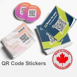 May include: A selection of QR code stickers with a variety of designs. The stickers are printed on white paper with a glossy finish. The stickers feature different colours and designs, including a pink and white sticker with the text "Studio 85" and a green and blue sticker with the text "Scan to pay".