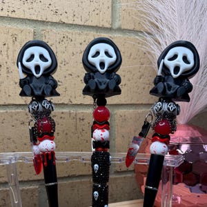May include: Three pens featuring a Ghostface character topper, black barrels, and decorative beads. The pens include red and white blood-splattered beads, small knife charms, and one pen barrel covered in black rhinestones with small Ghostface faces.